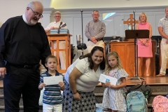Pastor Ken and Sarah Beth Wood smile with two young kids holding envelopes